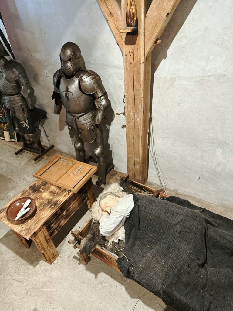 Suits of armor stand near a wooden bed with a sleeping figure inside