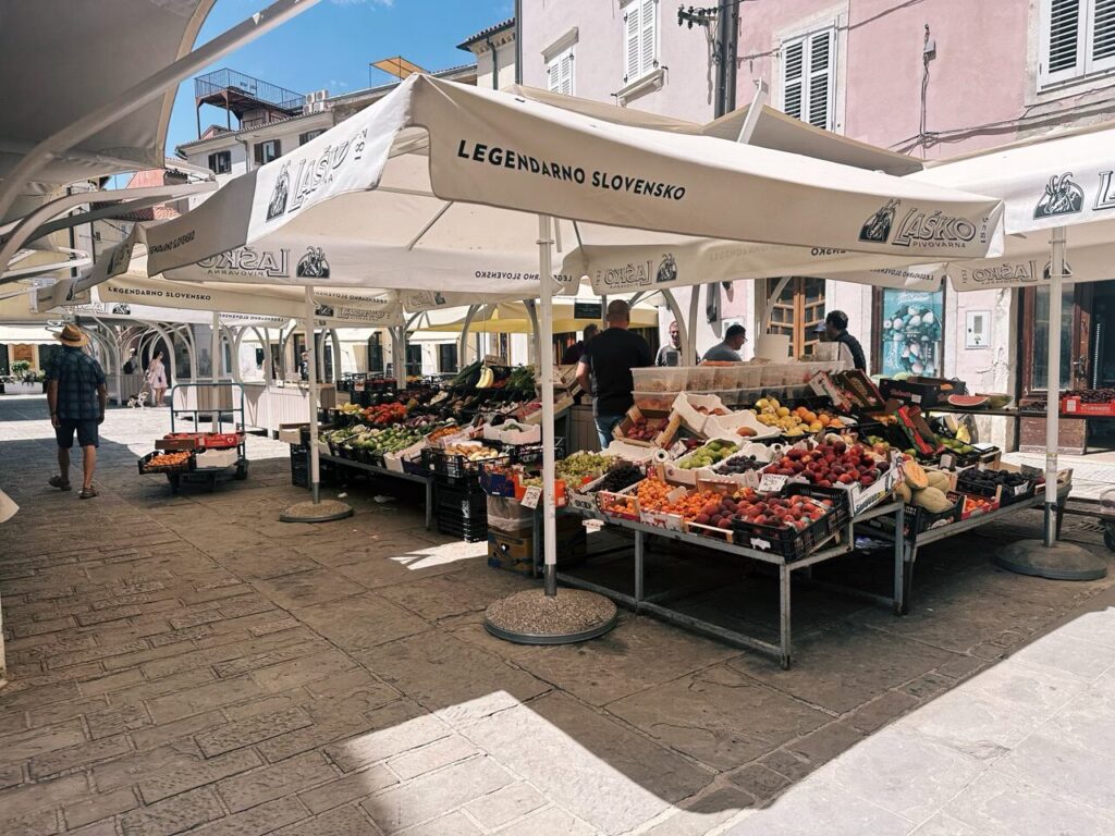 Outdoor fruit and vegetable market stalls under white umbrellas in Piran