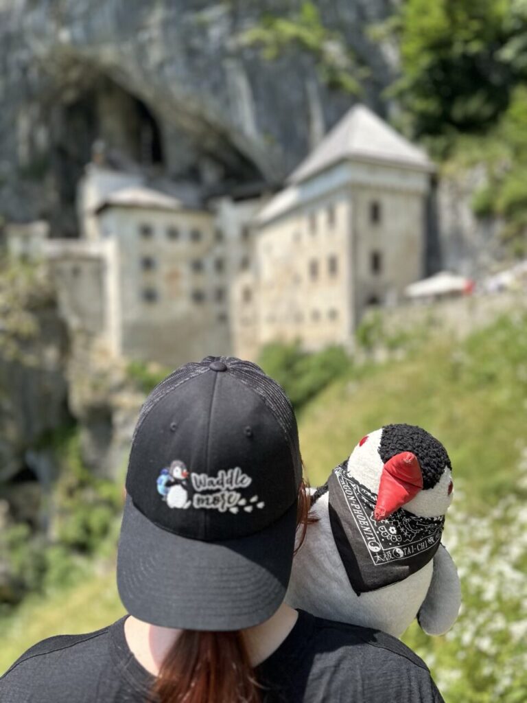 Person wearing a penguin logo baseball cap with Miško plush toy at Predjama Castle